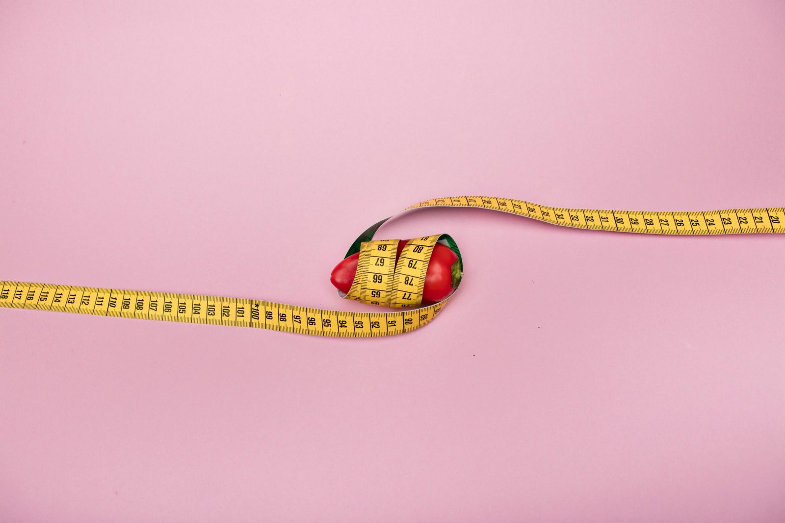 A red bell pepper wrapped with a yellow measuring tape on a pink background symbolizing diet and health.